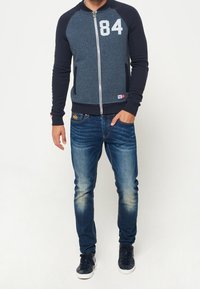 Marinfärgad zip-up jacka med melerat kropp, kontrasterande ärmar och stor vit tryck '84'. Kombinerad med ljusblå jeans och marinfärgade sneakers.