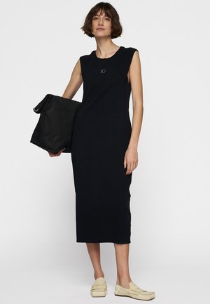 Abito midi senza maniche di colore blu navy con scollatura rotonda, caratterizzato da un piccolo logo. Il modello tiene una borsa nera e indossa scarpe slip-on di colore crema.