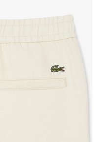 Off-white broekgedeelte met elastische tailleband, achterste paspelzak en klein groen krokodillenlogo geborduurd boven de zak.