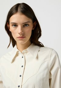 Camicia button-up bianca sporca con colletto a punta, bottoni blu e design in stile western sullo yoke. Tessuto liscio con una sottile texture.