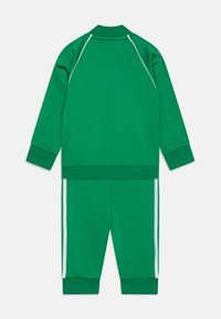 Fato atlético verde feito de material liso. Possui uma jaqueta com fecho éclair, colarinho e punhos canelados, e calças a condizer com riscas brancas nas laterais.