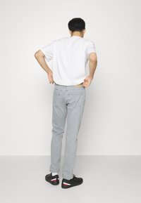 Jeans in denim grigio con taglio dritto, indossati con una t-shirt bianca e scarpe nere da ginnastica con dettagli rossi, bianchi e blu. Sfondo semplice.