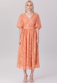 Robe en dentelle pêche avec un décolleté en V, des manches bouffantes et une taille ceinturée. La longueur midi présente un motif floral et une texture transparente.