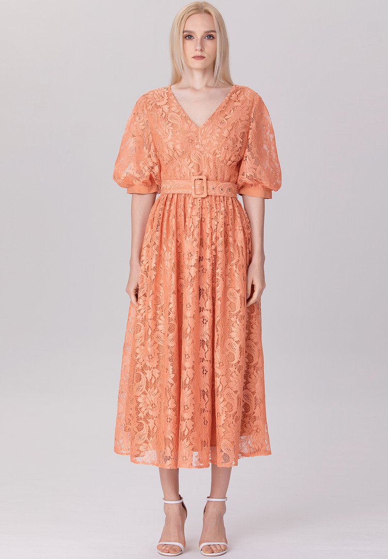 Robe en dentelle pêche avec un décolleté en V, des manches bouffantes et une taille ceinturée. La longueur midi présente un motif floral et une texture transparente.