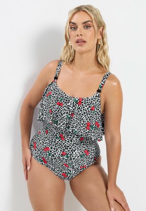 Lyshåret kvinde iført en sort-hvid leopardmønstret badedragt med røde kirsebærmotiver, poserende mod en hvid væg.