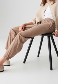 Femme portant un pantalon et une veste beige, assise sur un tabouret noir avec des chaussures claires sur un fond uni et clair.