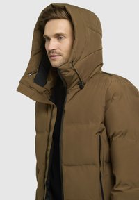 Puffjacka i mörkbrunt med hög krage och stor huva, med dragkedjefickor och snörningsdetaljer. Texturerad, isolerad design.