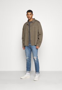 Casaco castanho com botões sobre uma camisa cinza, combinado com jeans rasgados de um azul claro e ténis brancos. Design minimalista, ajuste descontraído.