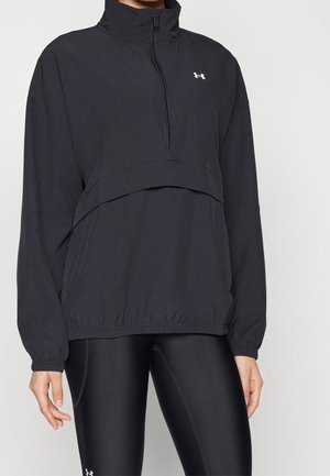 Personne portant un pullover noir à fermeture éclair quart, avec poignets et ourlet élastiques, associé à des leggings noirs ajustés ornés d'un petit logo blanc.
