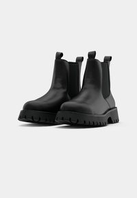 Mustad mustad Chelsea saapad elastsete külgpanelidega, ristkülikukujuliste tõmbevtikatega ja paksude soontega tallaosadega. Ükslane pind minimaalsete detailidega.