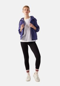 Paarse lichtgewicht jas, witte t-shirt, zwarte leggings en witte sneakers met rode en witte gestreepte sokken. Gladde stof en casual pasvorm.