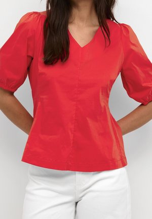 Femme portant un chemisier rouge à col en V avec des manches bouffantes et un pantalon blanc, debout les mains derrière le dos.