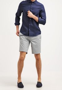 Marinblå skjorta med knappar och uppvikta ärmar, tillsammans med ljusgrå shorts. Modellen bär marinblå loafers. Texturerad tyg och skräddarsydd passform synlig.
