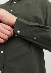 Tume roheline puuvillane särk nööpidega mansettides, ühe rinnataskuga ja valgete nuppudega. Kanga pind tundub pehme ja kerge tekstuuriga.