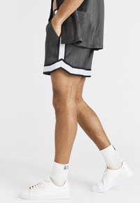 Shorts de sport noirs à motif géométrique, avec des bandes latérales blanches, accompagnés de baskets blanches et de chaussettes blanches côtelées.