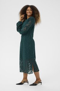 Lace long-sleeve dress in dark green, featuring floral patterns, a mid-length hem with side slits, paired with black heeled shoes.