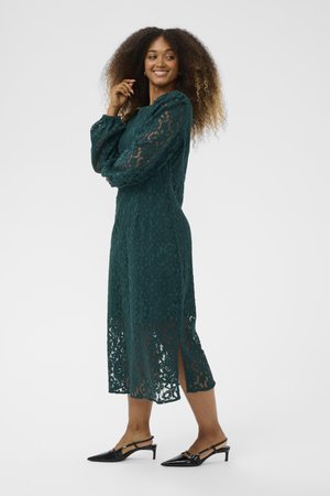Lace long-sleeve dress in dark green, featuring floral patterns, a mid-length hem with side slits, paired with black heeled shoes.