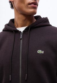 Κοντινό πλάνο ενός ατόμου που φοράει μαύρο φούτερ με φερμουάρ, με πράσινο λογότυπο κροκοδείλου στο στήθος και μαύρα κορδόνια.