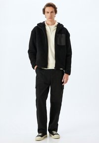 Chaqueta de forro polar negra con capucha, cierre de cremallera y un bolsillo lateral. Lleva puesta sobre una sudadera blanca y combinada con pantalones cargo negros y zapatos.