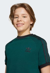 Žalias trumpas rankovės marškinėlis su juodais akcentais ir trimis juodomis juostelėmis ant pečių. Ant krūtinės yra juodai siuvinėtas adidas logotipas.