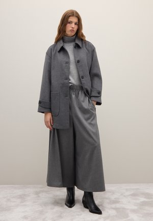 Mujer joven con un cuello alto gris, abrigo gris de gran tamaño, pantalones grises de pierna ancha y botas negras puntiagudas, de pie frente a un fondo liso.