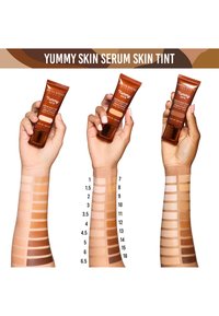 God smak Serum Skin Tint i en brun tub. Tre händer håller tuben och visar olika hudtoner på prov. Utställning av 16 numrerade nyanser.