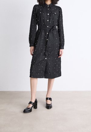 Nainen, jolla on päällään musta, pitkähihainen napitettava midimekko, jossa vyötärönauha ja niittikoristeet, yhdistettynä mustiin solkisorkkareihin.