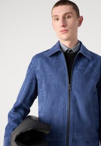 Μπλε σουέτ μπουφάν με φερμουάρ και κολάρο με περικοπές. Μαύρα δερμάτινα γάντια κρατημένα στον βραχίωνα. Διαθέτει ραμμένα ραφές και πλαϊνές τσέπες.