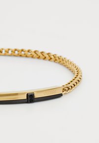 Guldtonet kædearmbånd med en sort rektangulær detalje gravert med "GUESS" og to små sorte sten på en hvid baggrund.