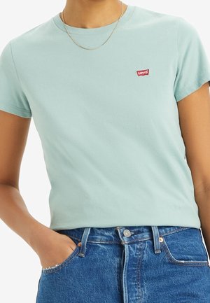 Osoba nosząca jasnoniebieską koszulkę Levi's i niebieskie jeansy, z jedną ręką w kieszeni i cienkim złotym naszyjnikiem na szyi.