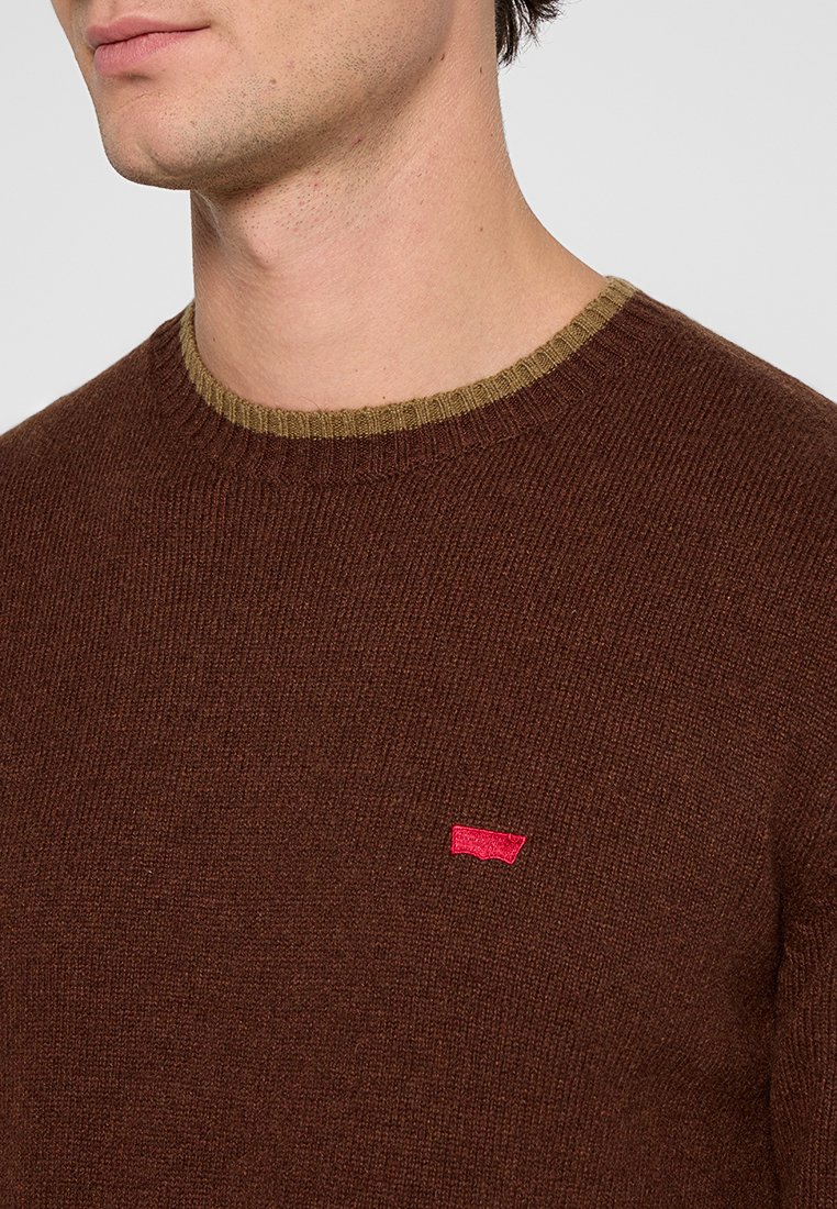 Brauner Strickpullover mit einem gerippten olivgrünen Kragen und kleinem rotem Logo auf der Brust. Der Stoff wirkt weich mit einer strukturierten Oberfläche.