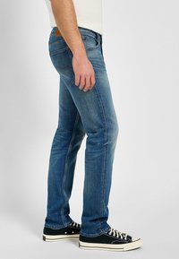 Vaqueros de mezclilla azul con un corte ajustado, un ligero desgaste y un diseño clásico de cinco bolsillos, acompañados de zapatillas altas negras.