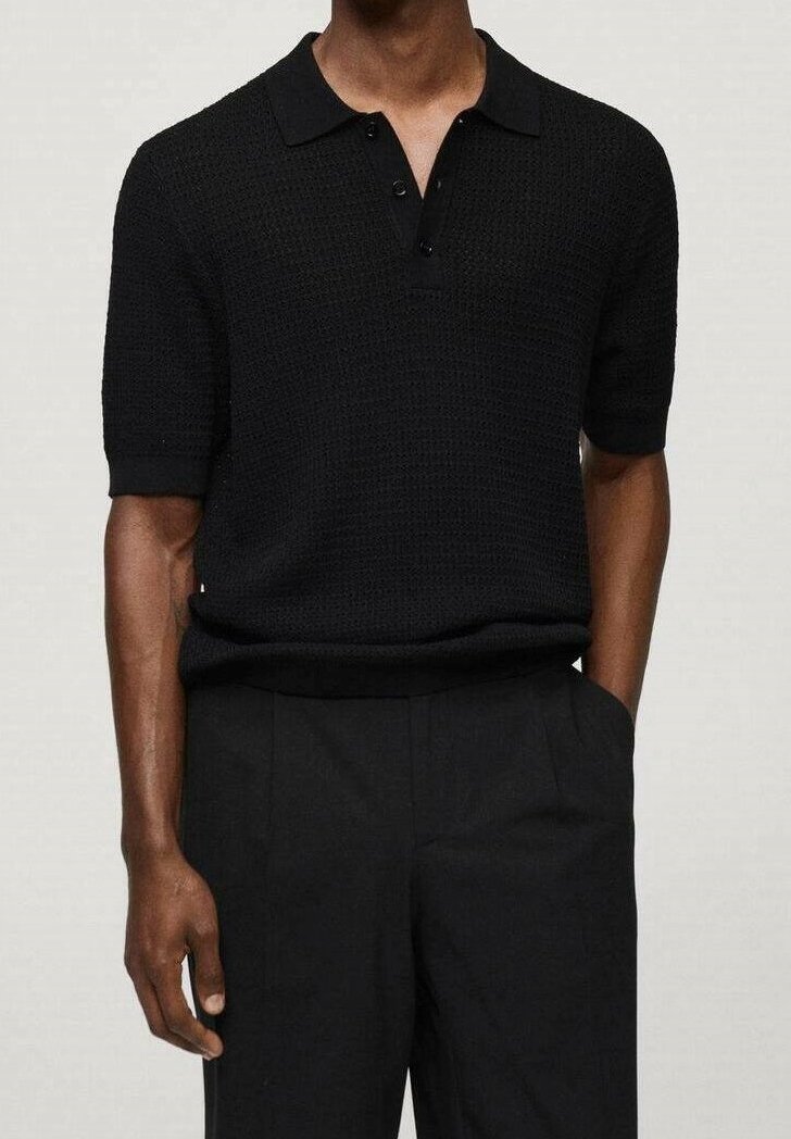 Homme portant un polo noir texturé à manches courtes, rentré dans un pantalon noir, une main dans la poche sur un fond clair.