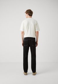 Hvit bomulls t-skjorte med korte ermer, kombinert med svarte jeans med kontrasterende hvit søm og lommer, sett fra baksiden.