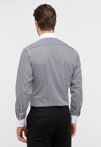 Maglietta a maniche lunghe con righe verticali bianche e nere, con colletto bianco e polsini con bottoni. Il tessuto appare liscio e aderente.