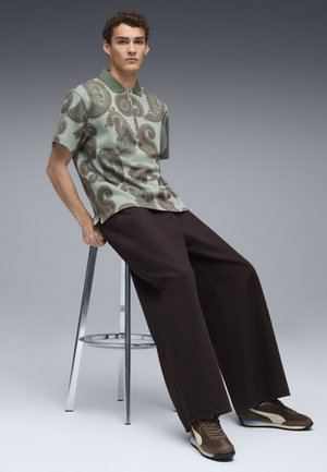 Camisa de manga corta con cuello en verde con estampado paisley; pantalones anchos marrones; zapatillas marrones con acentos azules; taburete metálico.