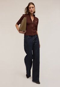 Top zip-up de culoare maro, cu garnituri verticale și mâneci scurte, asociat cu blugi evazați de culoare bleu închis cu buline albe și o geantă structurată de culoare maro.