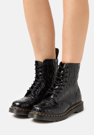 Bottes noires mi-mollet à lacets avec motif ondulé texturé et coutures jaunes portées sur des jambes nues sur fond blanc.