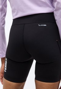 Κοντινό πλάνο ενός ατόμου που φοράει μαύρα αθλητικά σορτς με ψηλή μέση με τις ετικέτες "TERREX" και "CLIMACOOL", μαζί με ένα μακρυμάνικο τοπ σε λιλά χρώμα.