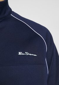 Navy blue high-neck pullover made from smooth fabric, featuring white piping along the shoulders and a small embroidered logo on the chest.