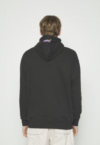 Personne portant un sweat à capuche noir avec le logo « puma » à l'arrière de la capuche, associé à un pantalon cargo blanc cassé, dos à la caméra.