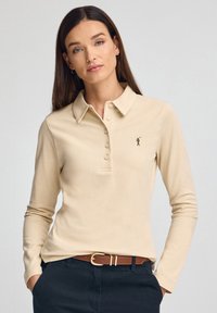 Polo de manga larga beige con cuello, cinco botones y pequeño logo marrón en el pecho izquierdo. Combinado con pantalones oscuros y cinturón marrón.