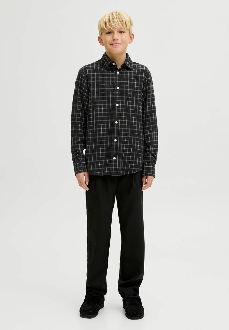 Melnā un baltā šaha raksta garpiedurkņu krekls ar pogājamo apkakli, valkāts kopā ar melnām biksēm un melnām kurpēm. Gluda auduma tekstūra.