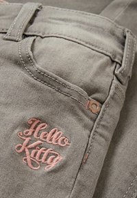 Gros plan sur une poche de jean gris avec le texte brodé "Hello Kitty" en rose et un petit rivet métallique en forme de fleur.