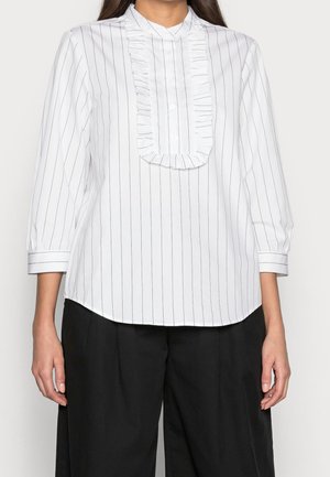 Blouse blanche avec fines rayures noires verticales, plastron froufroutant avec boutons, manches trois-quarts, associée à un pantalon noir ample.