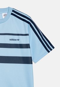 Camisa atlética de color azul claro con franjas horizontales de color azul oscuro, cuello redondo y el logo de Adidas en el área izquierda del pecho.