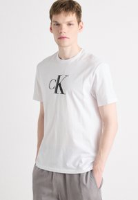 Balta medvilnės marškinėliai su ryškiu juodu "cK" logotipu krūtinėje. Dizainas apima trumpas rankoves ir apvalų kaklą.