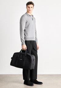 Pulover gri tricotat cu guler, geantă duffel din material negru cu mânere de sus, pantaloni negri și ghete închise la culoare pe un fundal neutru.