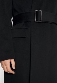 Abrigo de lana negro con cinturón de tela y hebilla metálica rectangular, mostrando la mano izquierda y un bolsillo de solapa en el lado izquierdo.