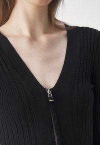 Body noir côtelé avec un décolleté en V profond et une ouverture frontale zippée métallique, présentant une texture en maille lisse.